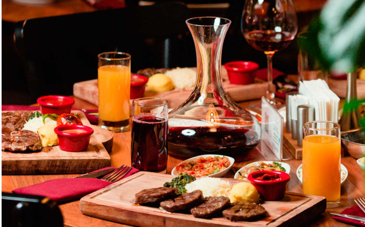 Uma mesa de jantar posta, em um restaurante, com um decanter de vinho tinto, dois copos com suco de laranja, tabua de carnes e acompanhamentos.