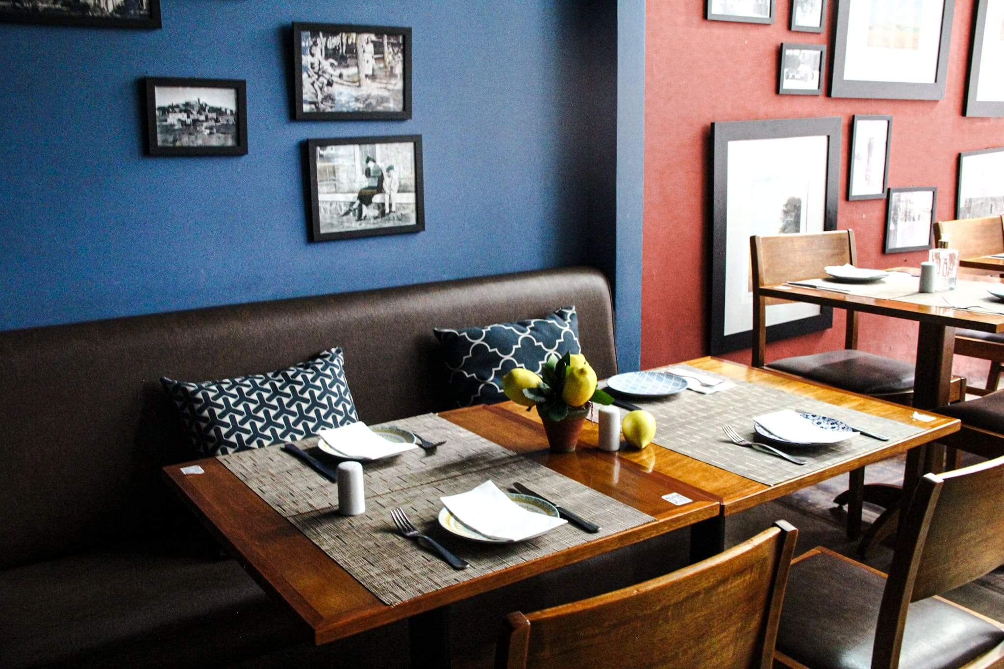 Foto mostra o ambiente interno do restaurante, com a parede sendo metade vermelha/metade azul. Sofá de couro com almofadas azuis encostados na parede azul de frente com mesas de madeira. Nas paredes há vários quadros em preto e branco.