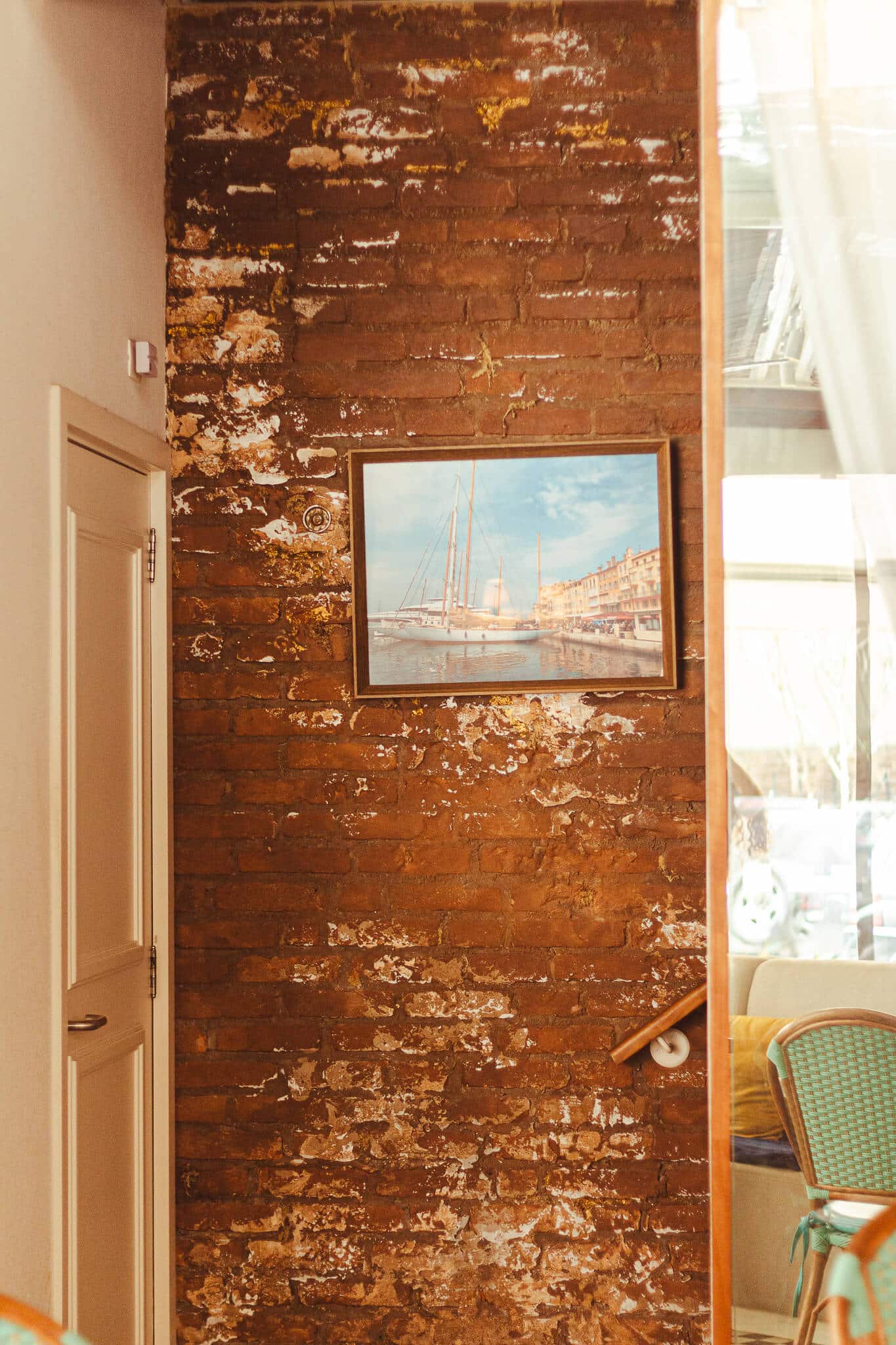 Ambiente interno do restaurante mostra uma parede rústica com um quadro pequeno que ocupa três palmos da parece. O quadro mostra uma arte mediterranea, com paisagem à beira-mar