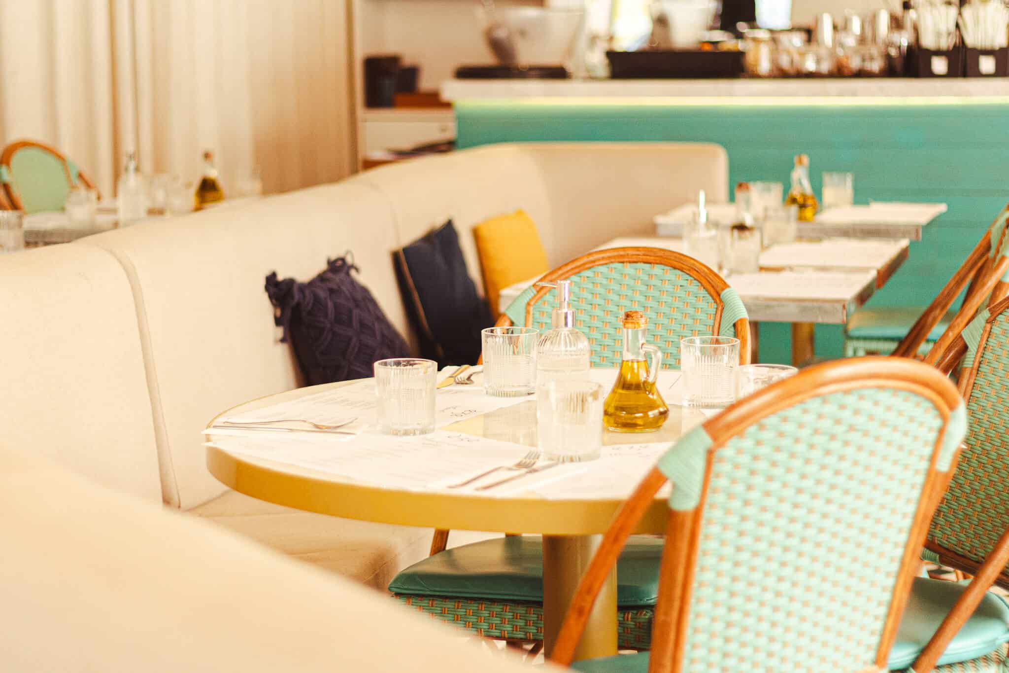 A foto mostra o ambiente interno do restaurante èze, com iluminação natural, sofá na cor clara em harmonizando com mesas redondas e, ao lado, mesas quadradas. A foto mostra duas cadeiras vazadas de forma simétrica simulando uma esteira de palha na cor azul, com bordas de madeira na cor clara. No sofá há 2 almofadas azuis e 1 amarela. Na mesa há copos vazios e uma azeiteira.