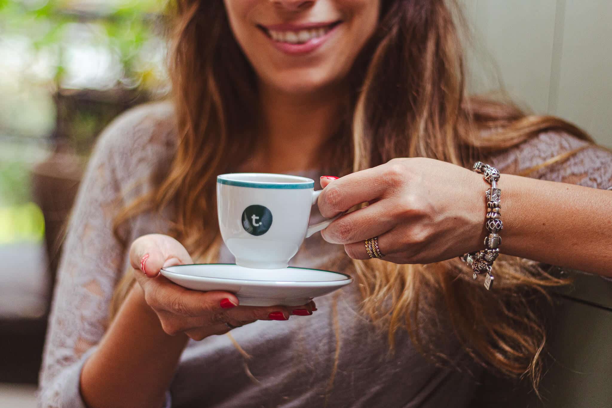 a foto mostra uma mulher branca, jovem, de cabelos longos, segurando uma xícara e pires com café expresso. a xícara contem o logo do tea connection e a mulher está sorrindo.