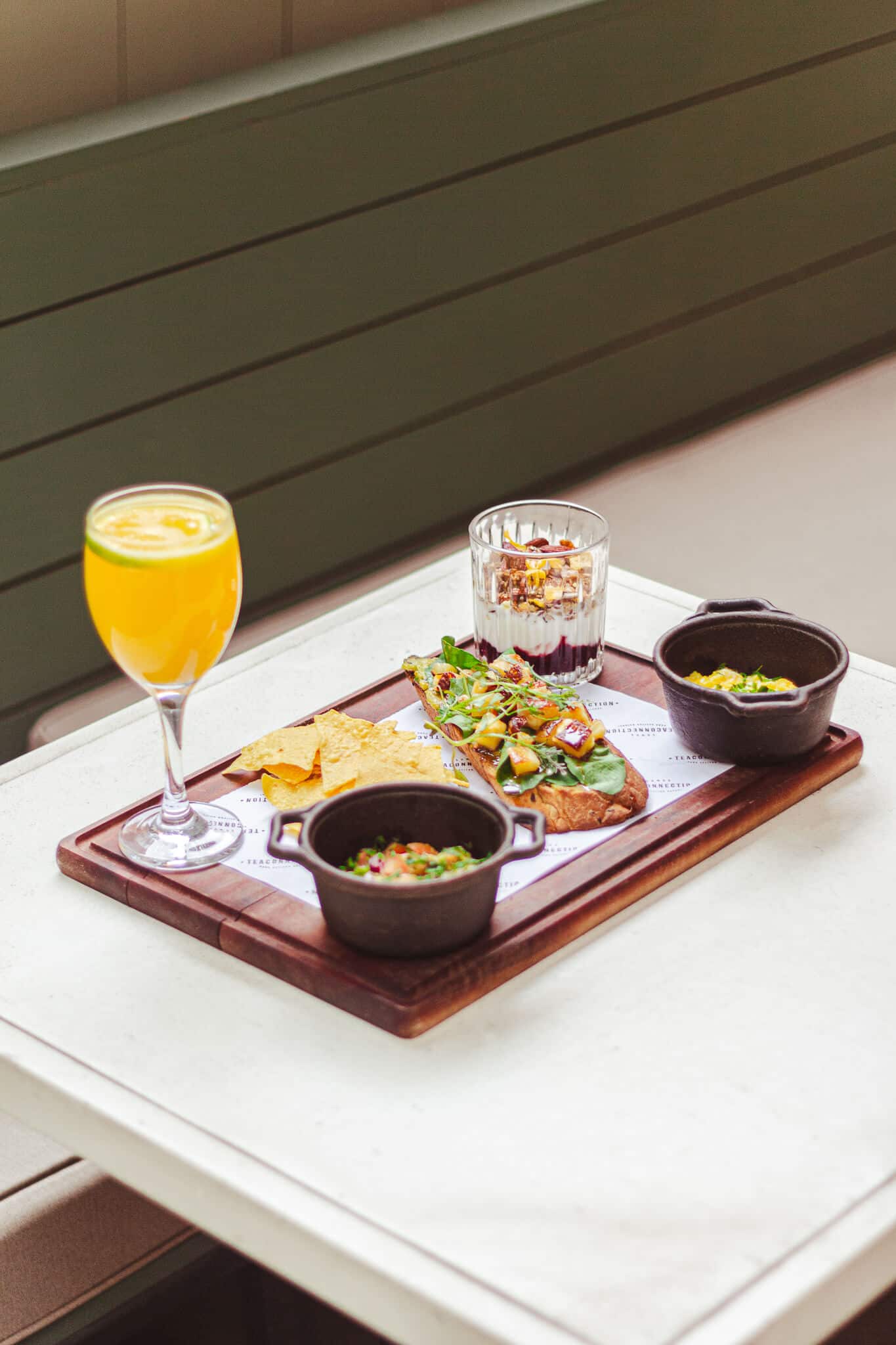 foto mostra uma mesa ao livre, sendo a mesa branca com uma bandeja marrom em cima. na bandeja há o brunch, com 1 toast de avocado, tigelas pretas com ovo mexidos, um copo de vidro com iogurte com frutas vermelhas e granola, além de uma taça cheia de suco de laranja.