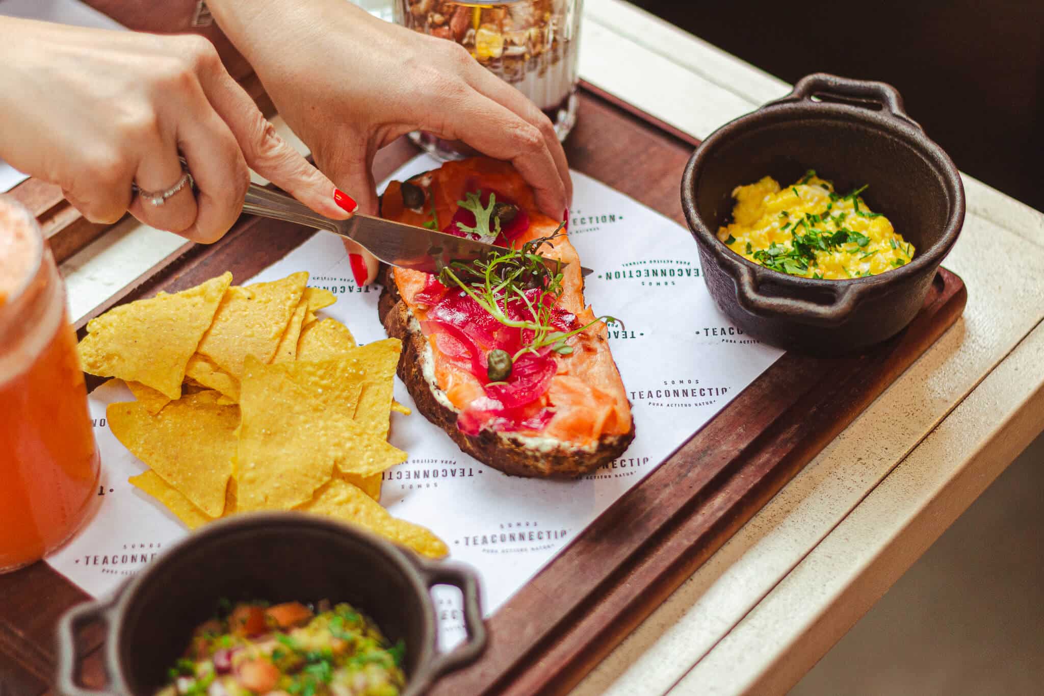 a foto mostra uma bandeja marrom em cima de uma mesa clara. na bandeja há potes pretos com ovos mexidos, nachos soltos e uma taça de mimosa. Uma pessoa branca está cortando com uma faca um toast de salmão ao meio.