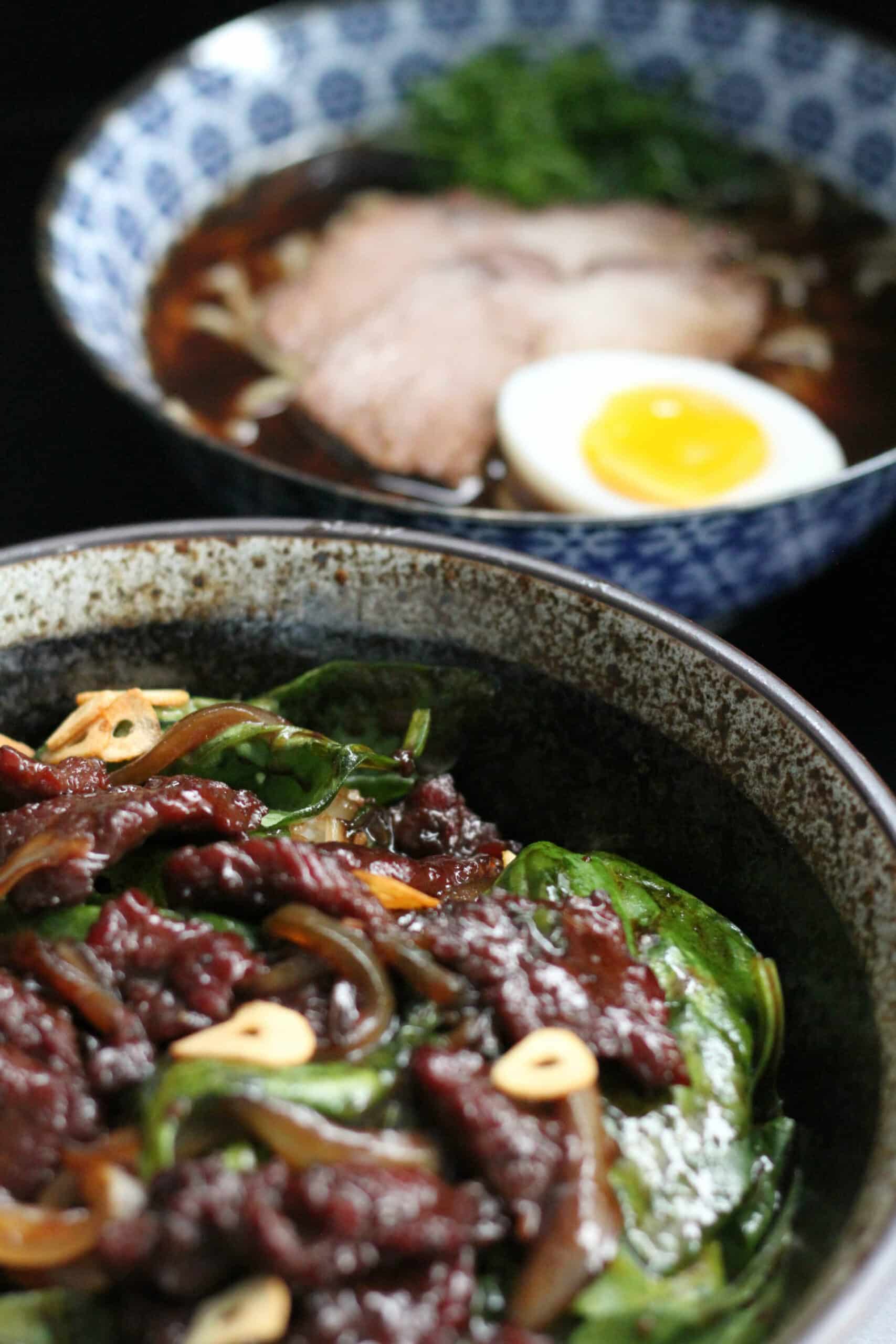 foto possui dois bowls em cima de uma mesa preta, em um deles contem o prato beef wok e no outro contem lamen. beef wok: Finas fatias de carne bovina salteadas com cebola, espinafre, alho frito, servido sobre arroz. ramen: Macarrão com caldo saboroso de porco e frango a base de shoyu, lombo suíno assado, ovo marinado e cebolinha