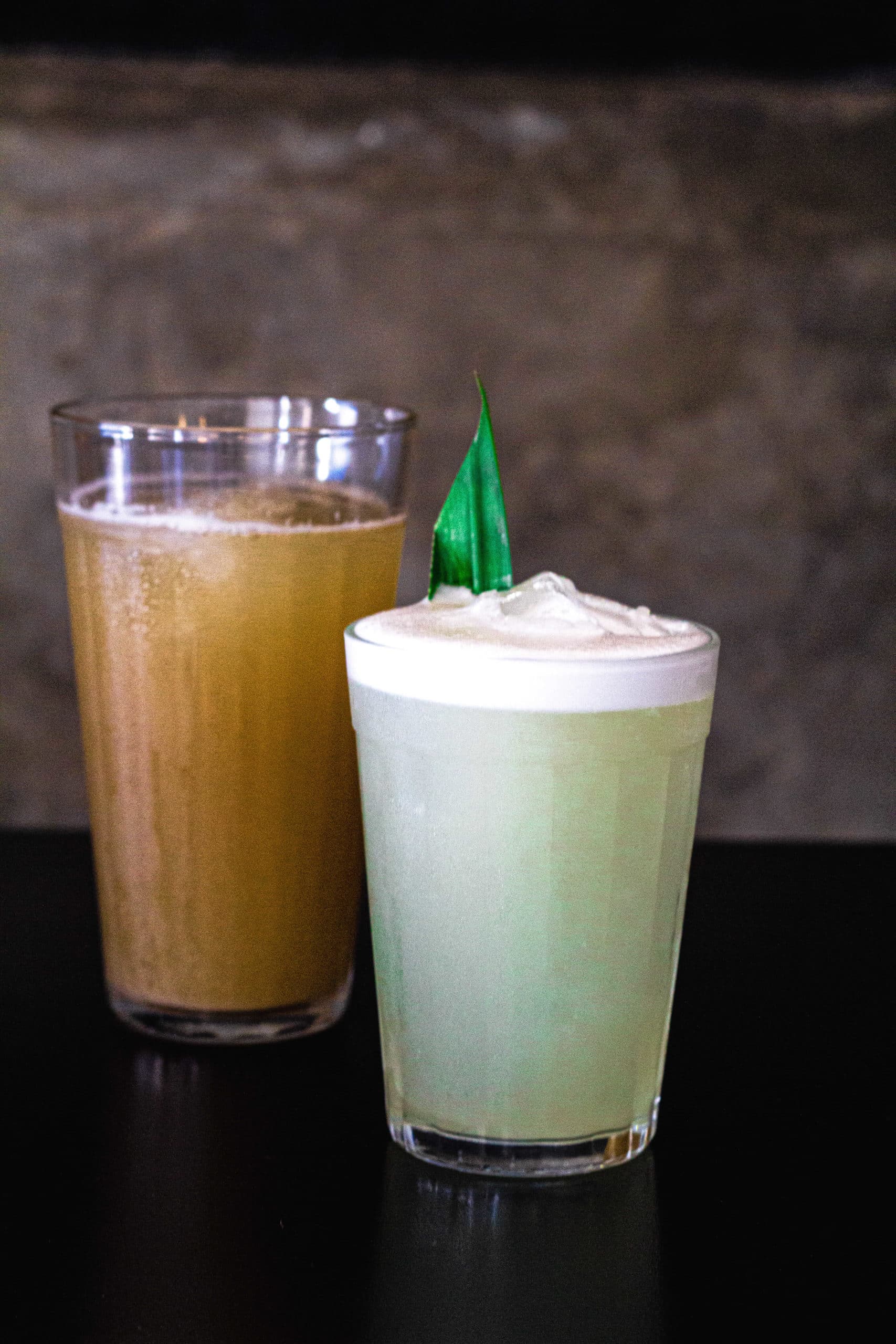 a foto mostra dois copos de drinks no Jui. Atrás um copo maior com refrigerante de gengibre de cor alaranjada. O outro copo menor é um drink de cor mais esverdeada.