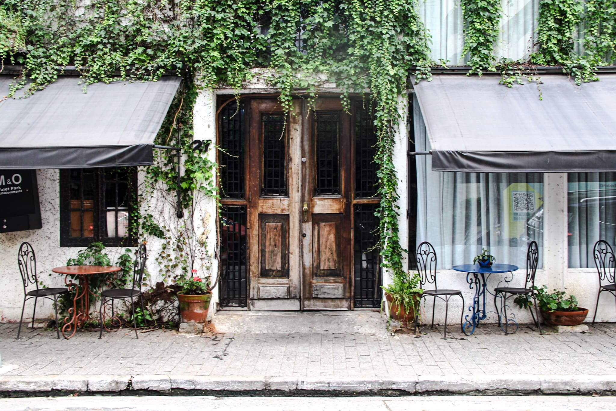 Foto mostra a fachada do restaurante Pina com plantas exuberantes, proporcionando um cenário fresco e acolhedor. Na foto mostra o parklet florido e, a porta no meio, é de madeira no estilo rústico.