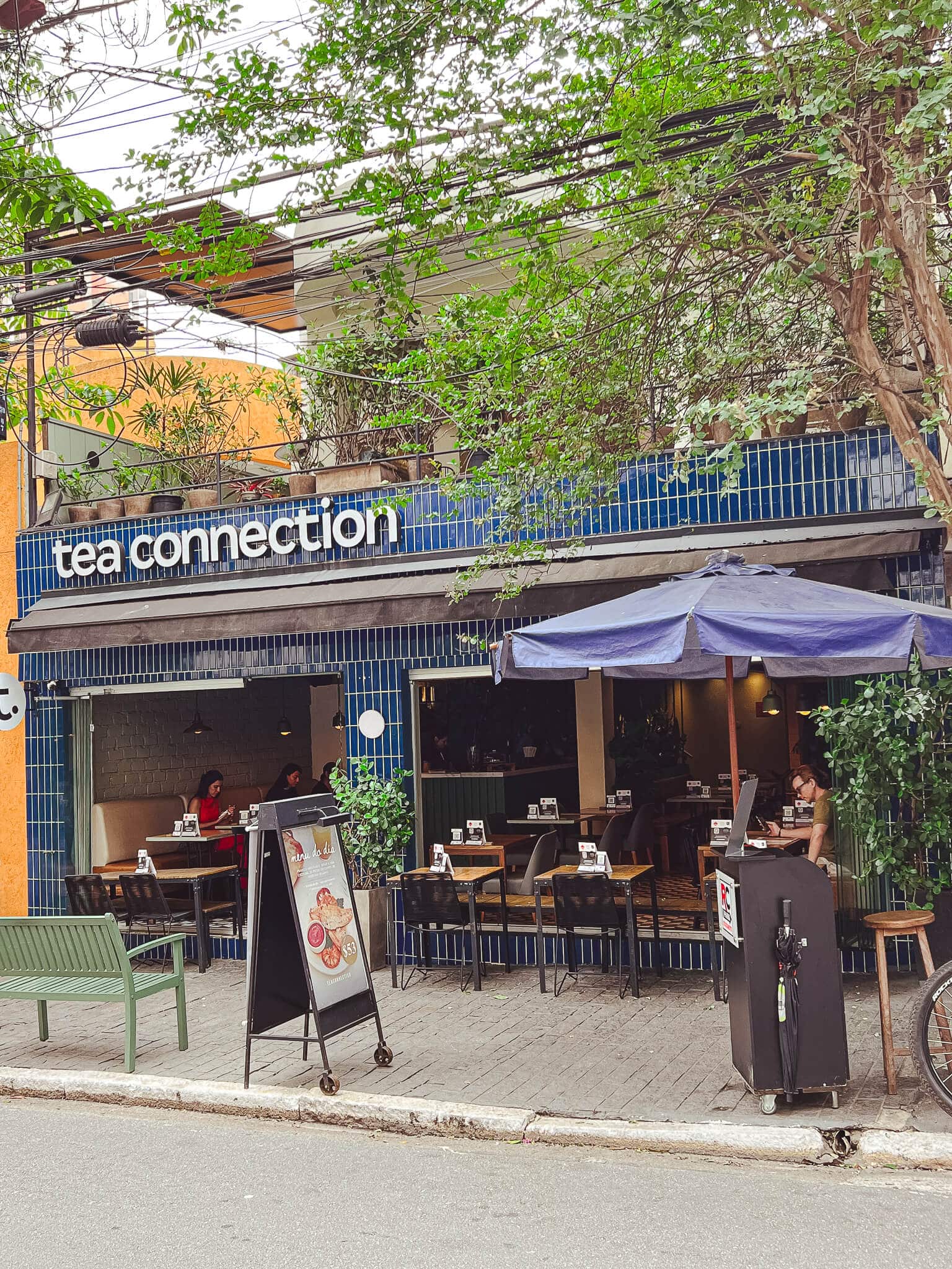 foto mostra a fachada do tea connection na unidade jardins. a rua é arborizada, há mesas pretas na parte de fora/calçada. o logo tea connection é escrito em letra minuscula e seu fundo é composto de azulejos azuis.