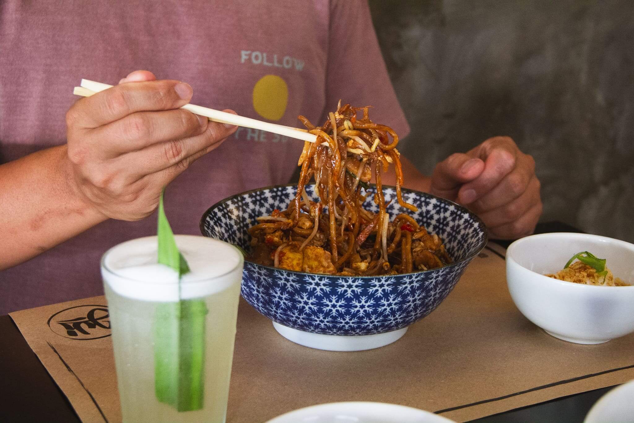 foto mostra uma pessoa com hashi comendo do mee goreng. ao lado há o drink nanas sour/stormy. com abacaxi e erva aromática pandan.