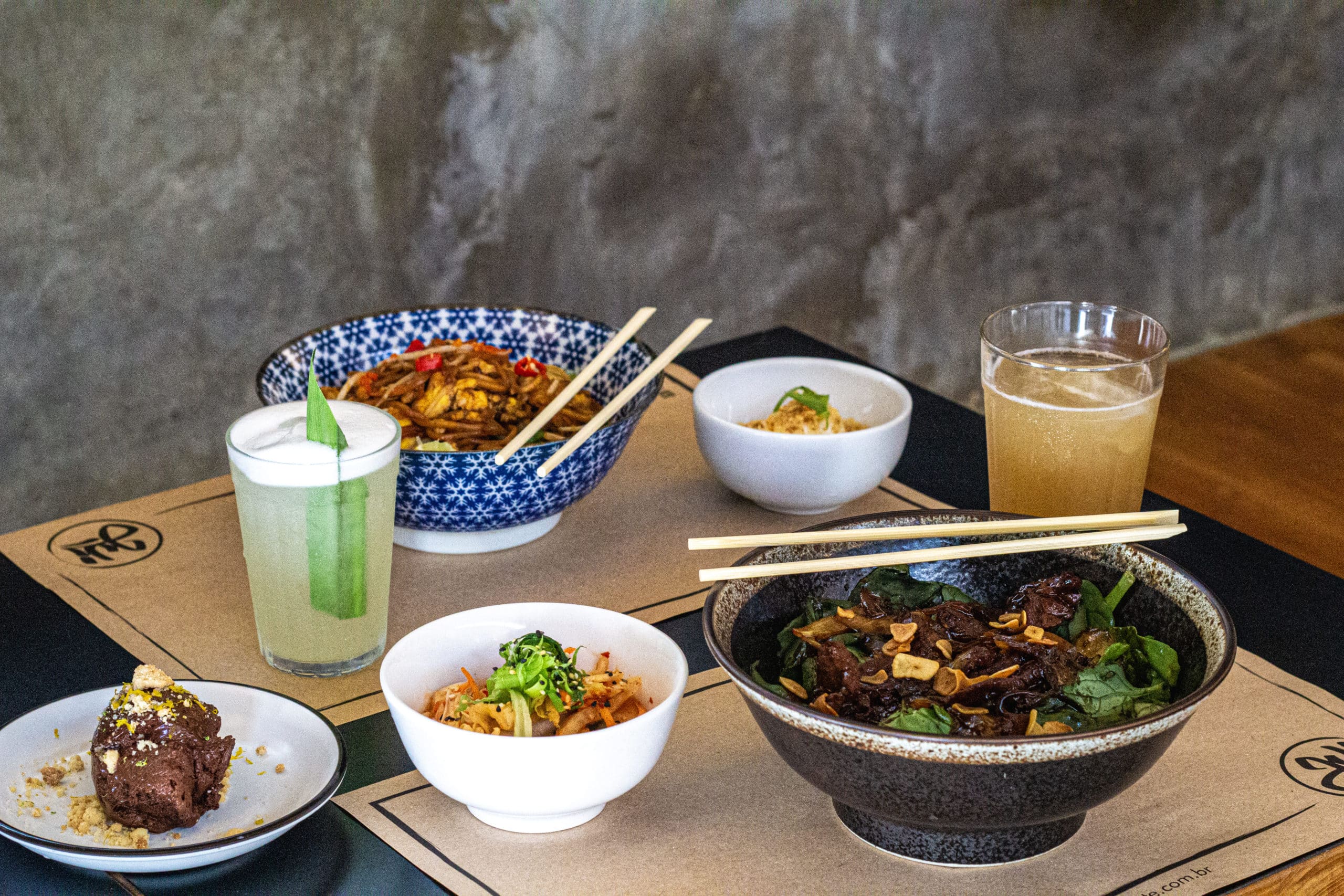Foto mostra uma mesa posta com todos os pratos, entradas e drinks descritos. Mee goreng, Beef wok, kimchi, ovo, ginger ale, nanas sour/stormy e, lado, a sobremesa.