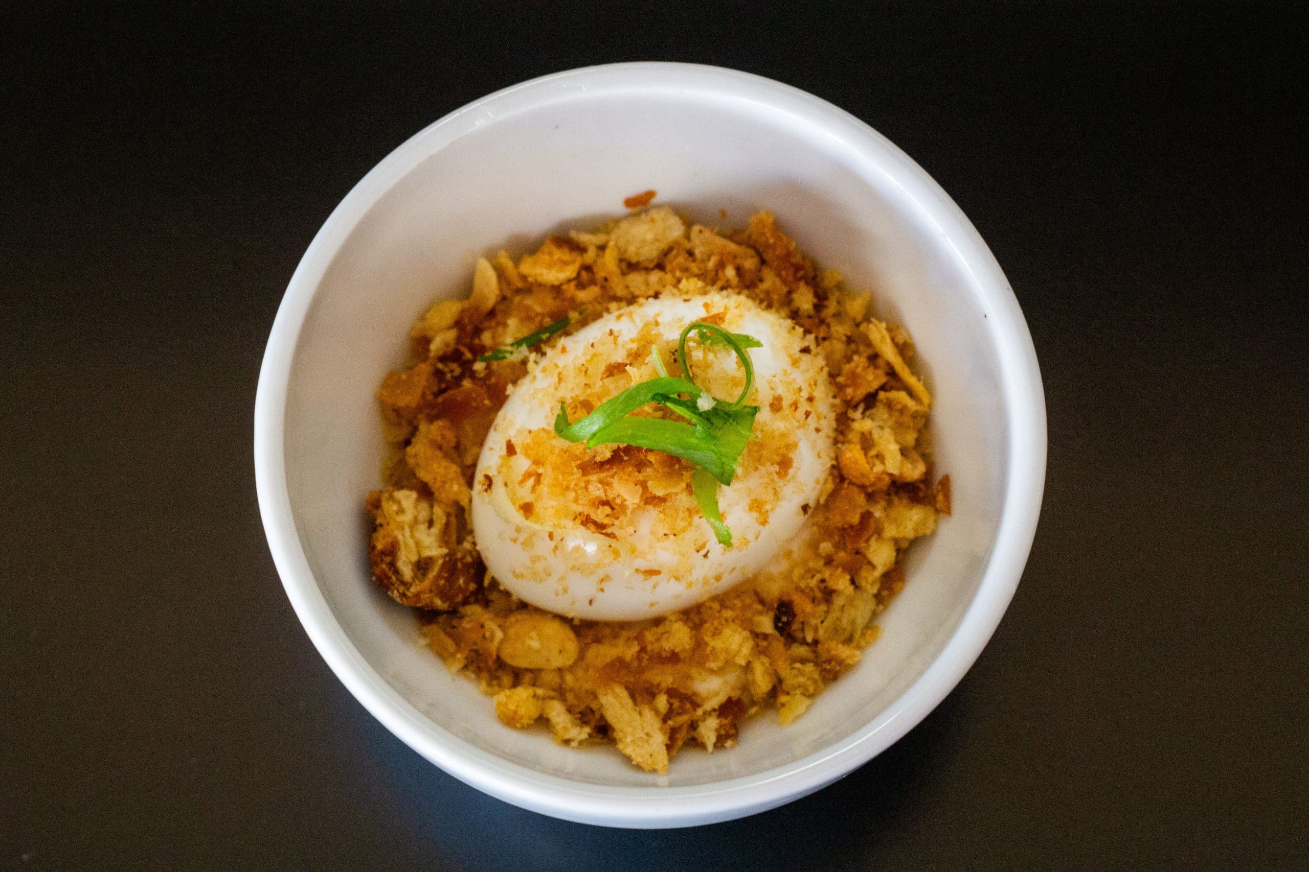 A foto mostra um bowl pequeno de porcelana branco, com a famosa entrada do Jui. O Ovo. ovo com gema mole, crocante de amendoim e creme de missô