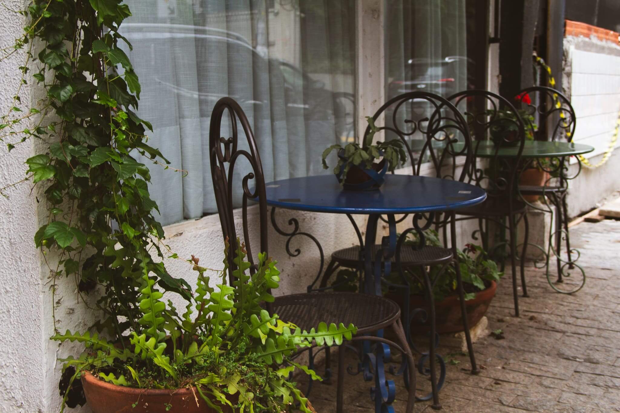 A foto mostra o lado de fora do Pina, com um parklet florido. Dupla de mesas e cadeiras elegantes e rústicas, com várias plantas e flores ao lado. A calçada é de paralelepípedo.