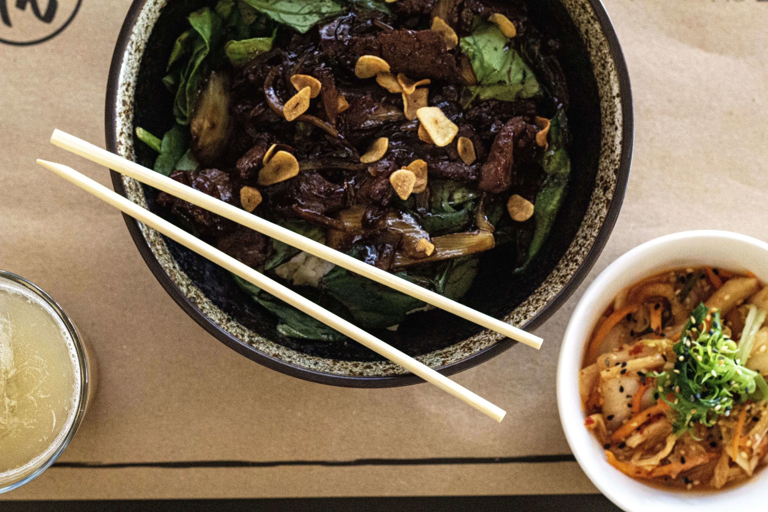 Foto contem um bowl principal com beef wok e hashis por cima. Ao lado há um bowl pequeno com kimchi (Acelga fermentada coreana). Beef wok contem: Finas fatias de carne bovina salteadas com cebola, espinafre, alho frito, servido sobre arroz.