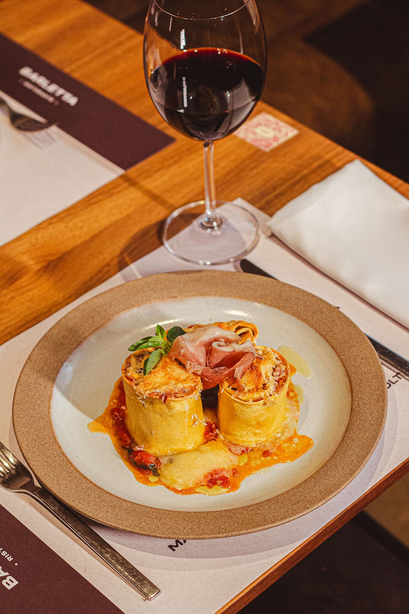 foto mostra uma mesa posta com o prato coliseu ao lado de uma taça de vinho. O coliseu é uma massa fresca e artesanal enrolada com presunto Culatello e Mussarela em uma cama de molho pomodoro, tudo isso gratinado ao forno.