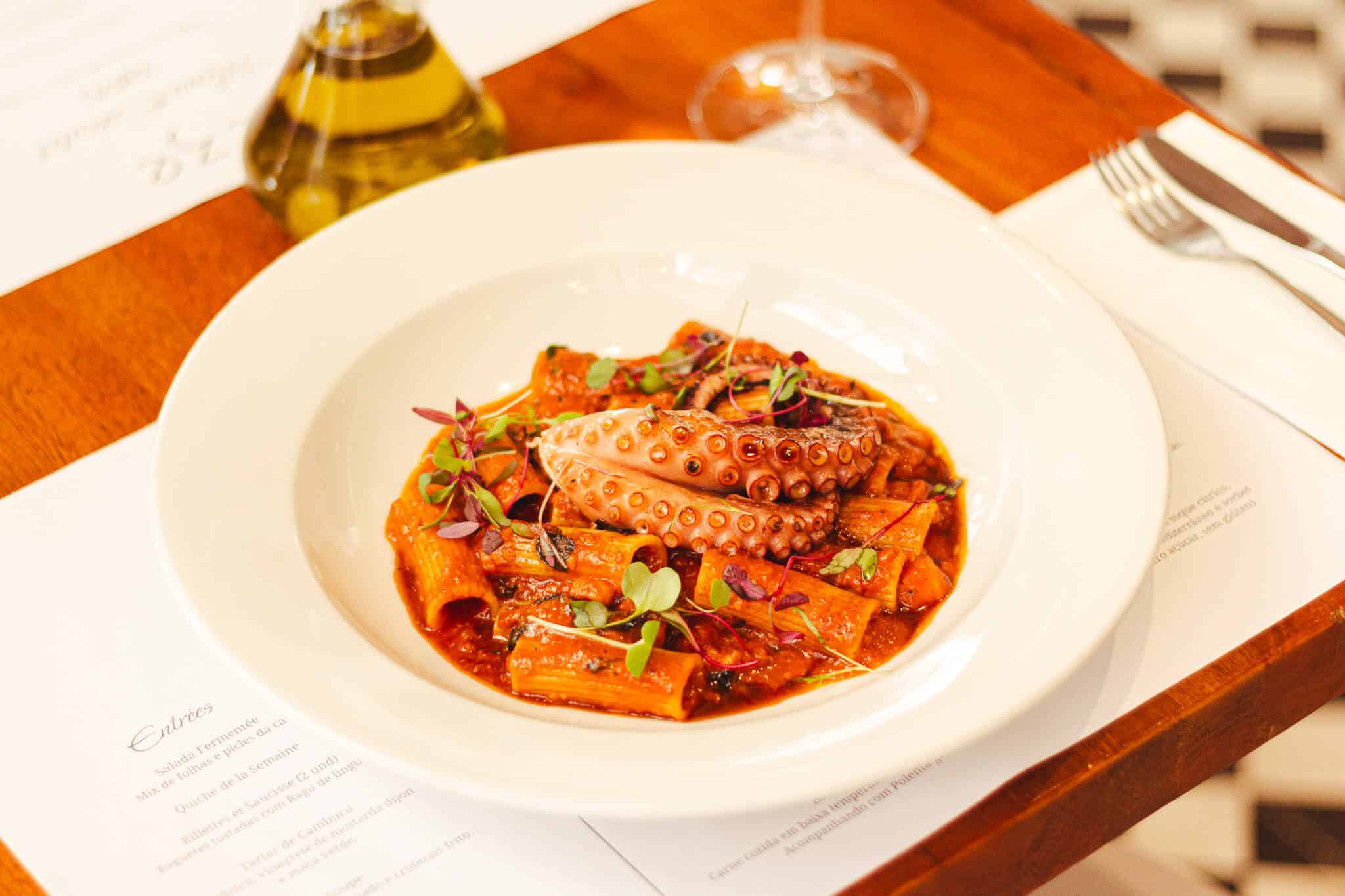 Foto mostra uma mesa posta com um prato fundo branco, dentro do prato há uma refeição com polvo com os seguintes ingredientes: Rigatoni, ragu de porco, pancetta curada, molho glacê e polvo grelhado na brasa.