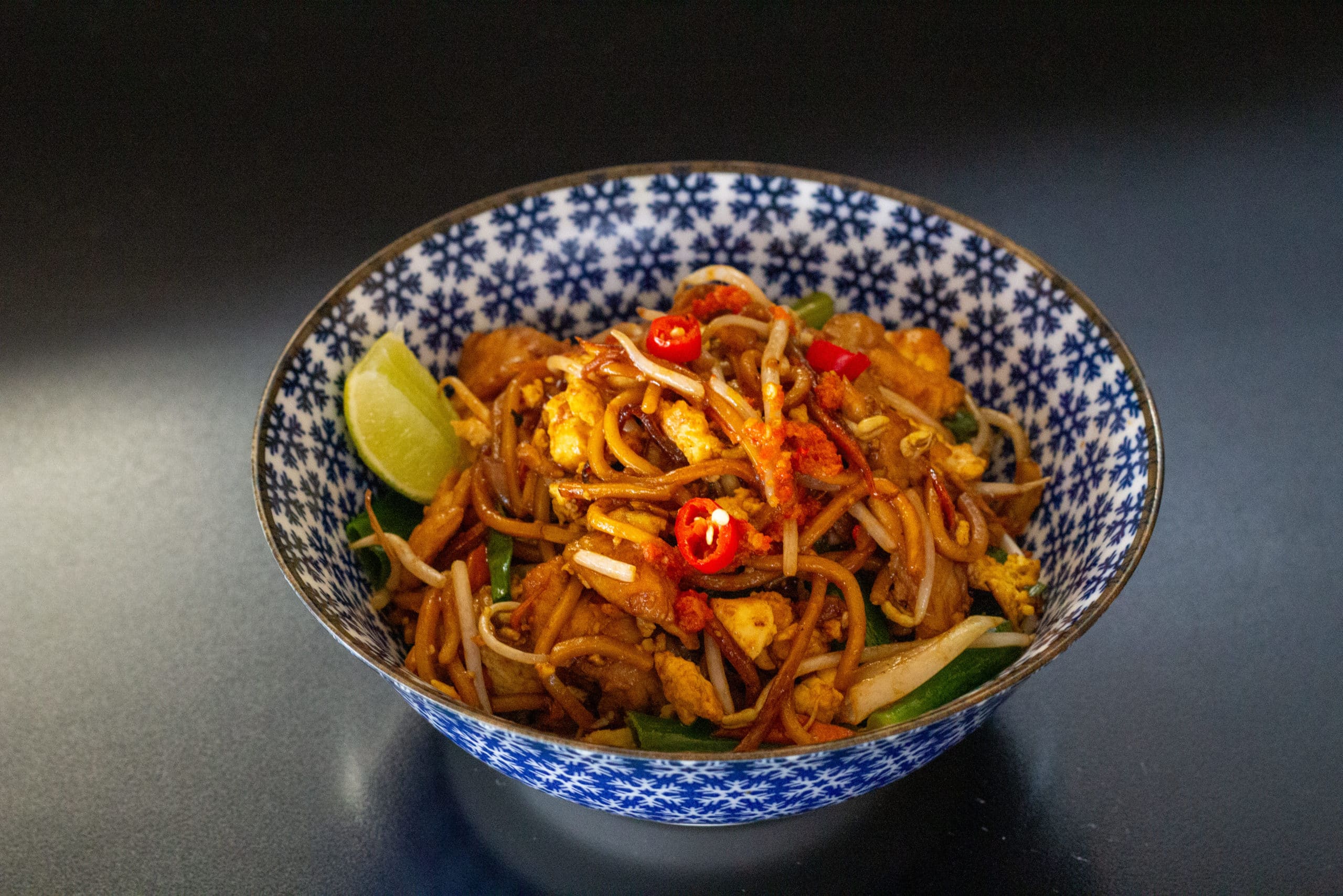 foto mostra um bowl de cerâmica com desenhos abstratos na cor azul. o bowl tem mee goreng. Macarrão frito, frango, ovo, cebola, cenoura, cebolinha e molho de pimenta 