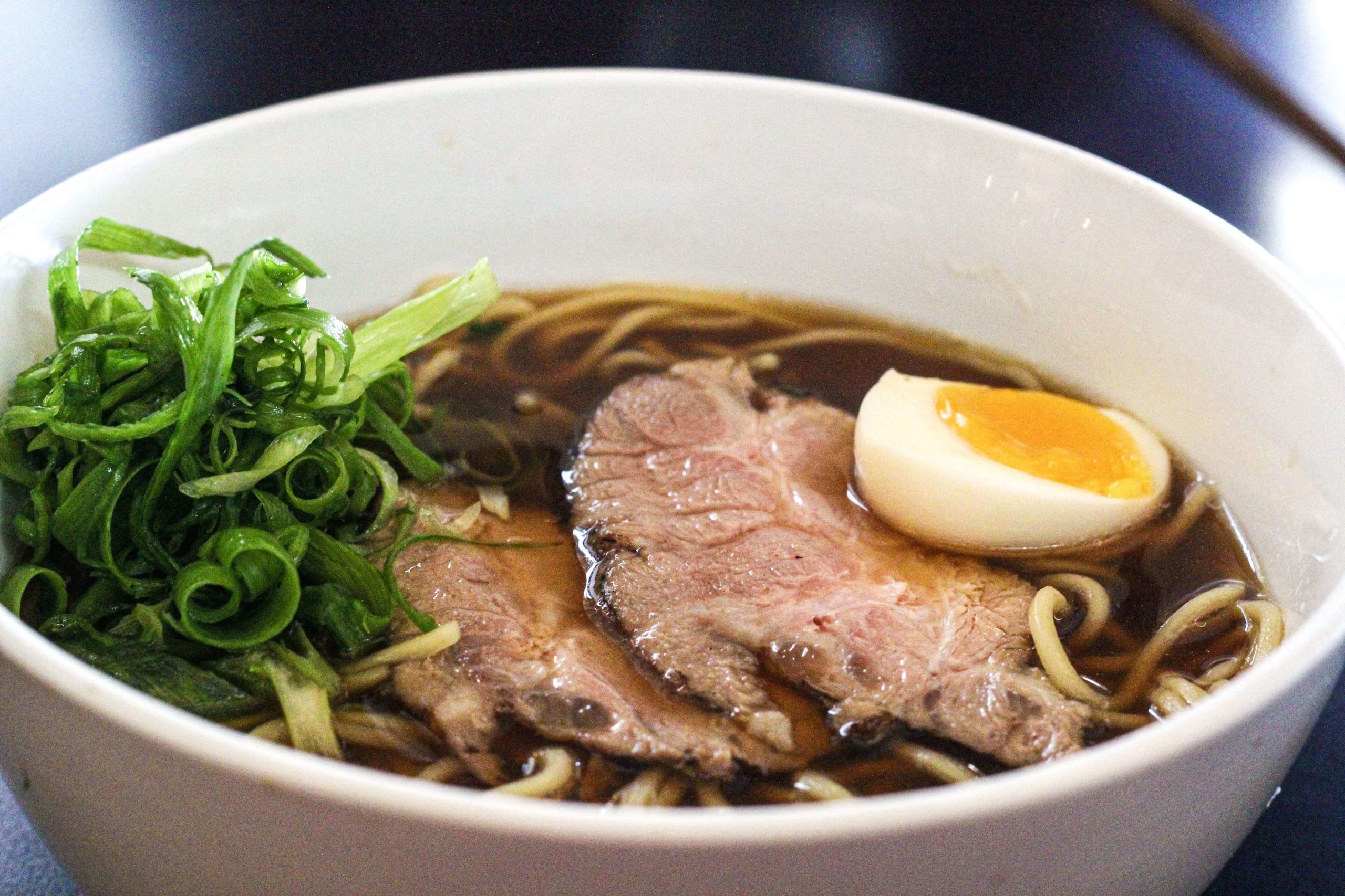 Foto mostra um bowl branco com ramen. Macarrão com caldo saboroso de porco e frango a base de shoyu, lombo suíno assado, ovo marinado e cebolinha