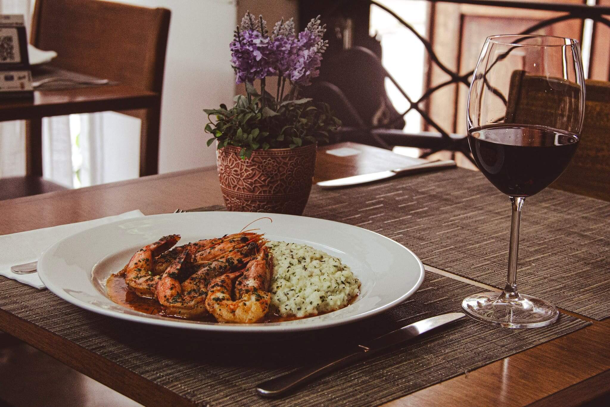 A foto mostra um prato branco com um risoto com ervas, parmesão e camarão rosa flambado com conhaque. A mesa está posta e possui um vaso de flor ao lado esquerdo e uma taça de vinho pela metade no lado direito.