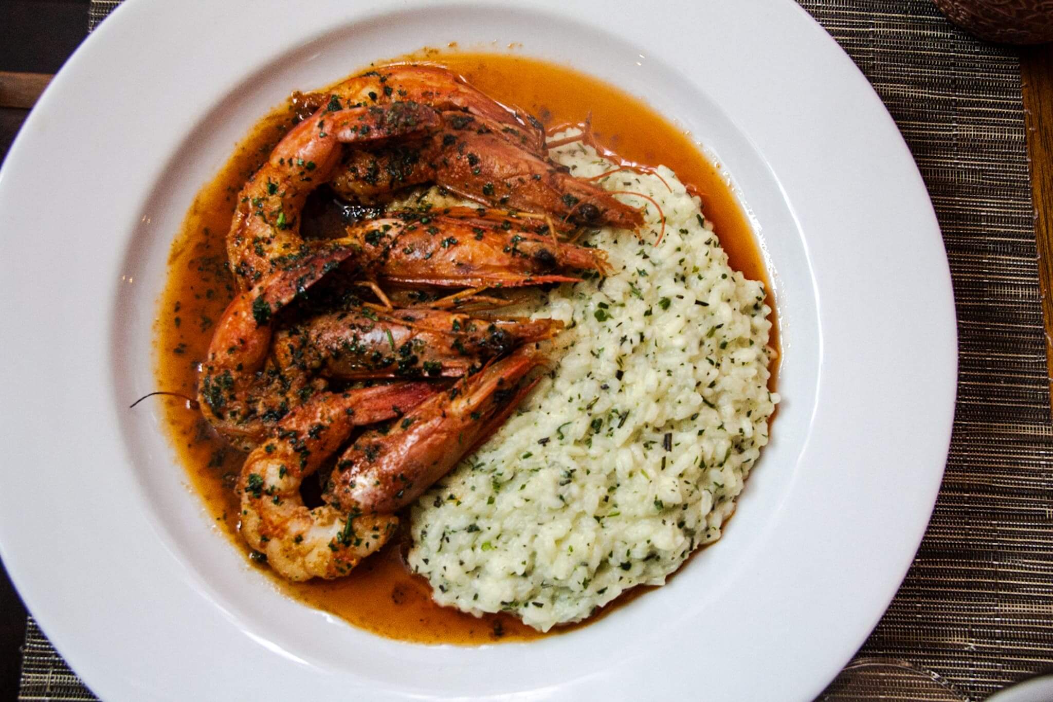 A foto mostra um prato branco com um risoto com ervas, parmesão e camarão rosa flambado com conhaque.