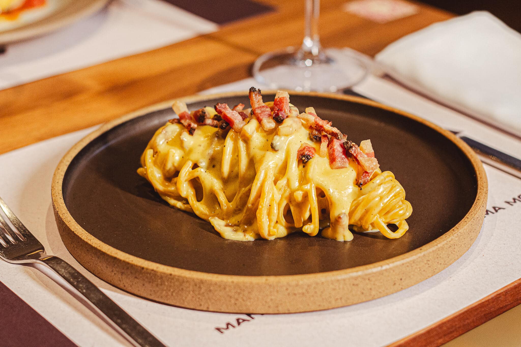 foto mostra um spaghetti em uma mesa posta, com prato redondo. Spaghetti alla chitarra Carbonara, com Guanciale (Bochecha de porco), gema de ovos caipiras, Pecorino e pimenta do reino! 