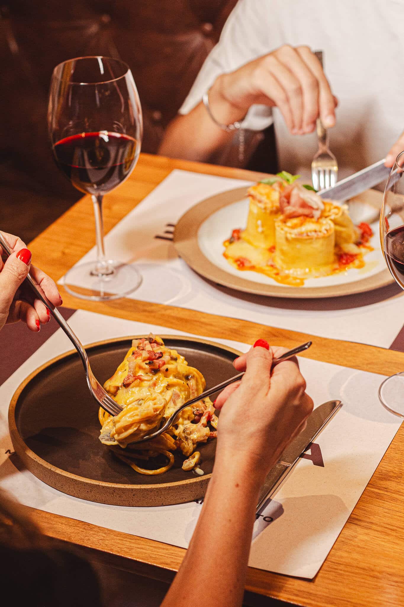 foto mostra duas pessoas comendo à mesa posta, uma com coliseu, a outra com spaghetti. há uma taça de vinho na mesa