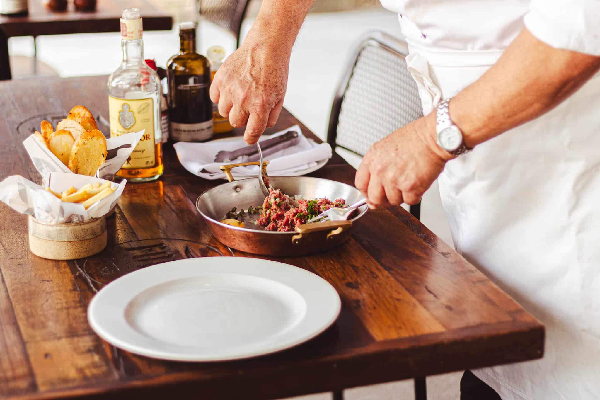 foto mostra o processo de preparação do steak tartare direto na mesa, o chef está com as mãos na panela fazendo a mistura dos ingredientes