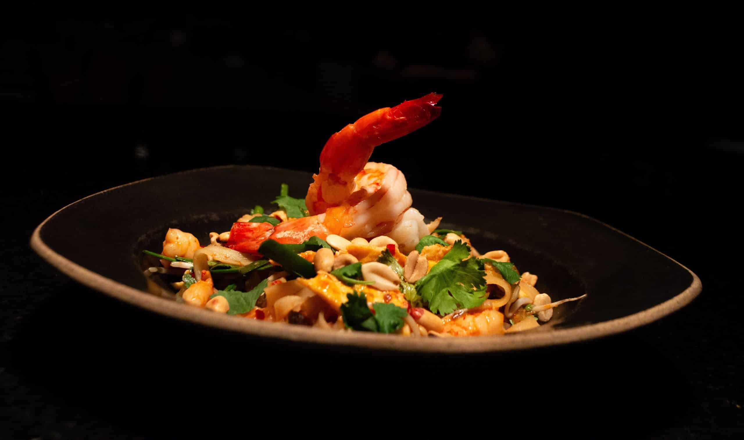 foto mostra um pad thai em um prato fundo preto. o pad thai é um apimentado talharim de arroz que acompanha ovo, camarão, broto de feijão, pimenta, coentro, amendoim e especiarias!