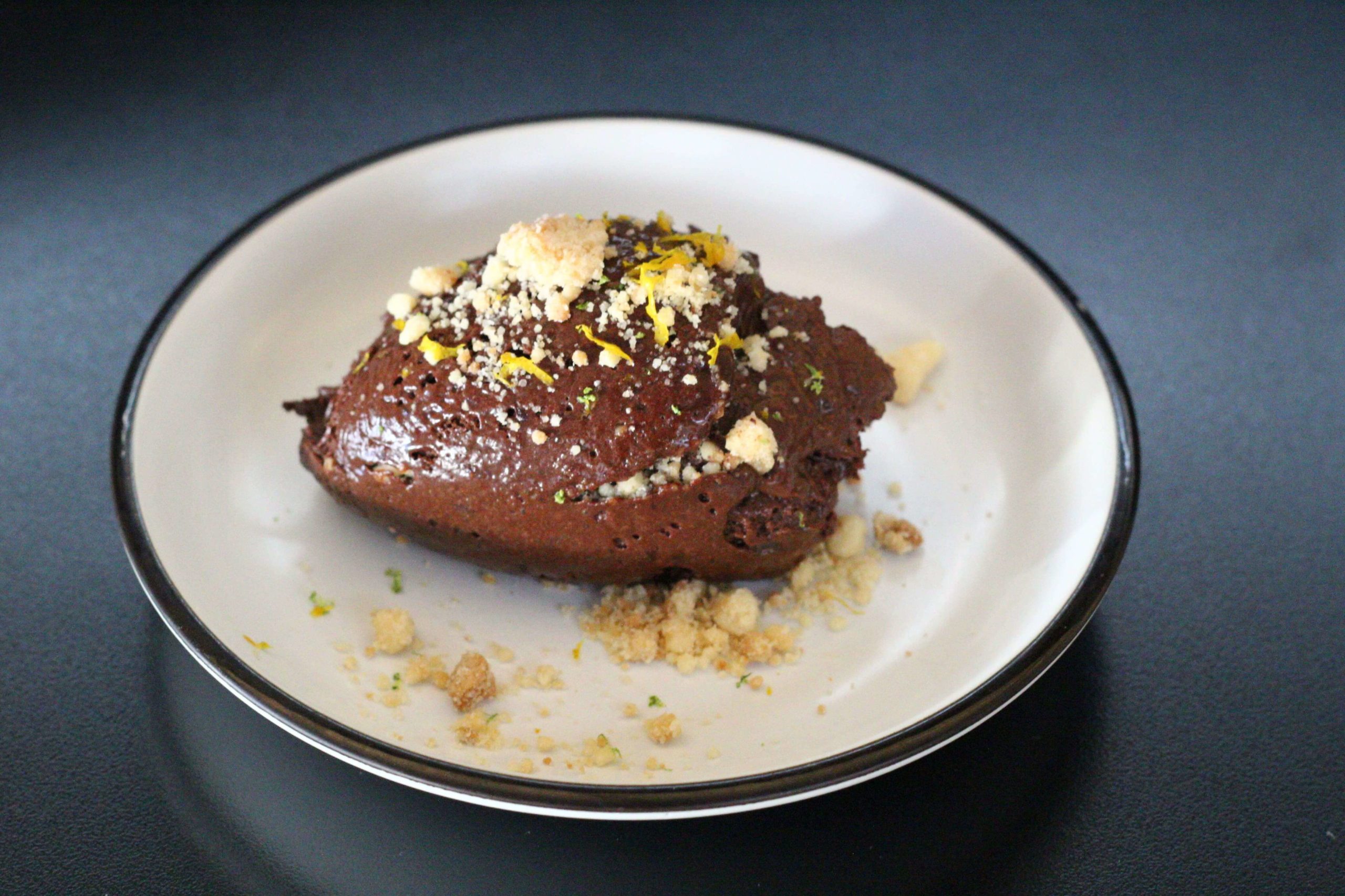 Foto contem um prato pequeno branco com listra circular na borda na cor preta. No prato há um mousse de chocolate. Mousse de Chocolate, com Laranja e Crumble