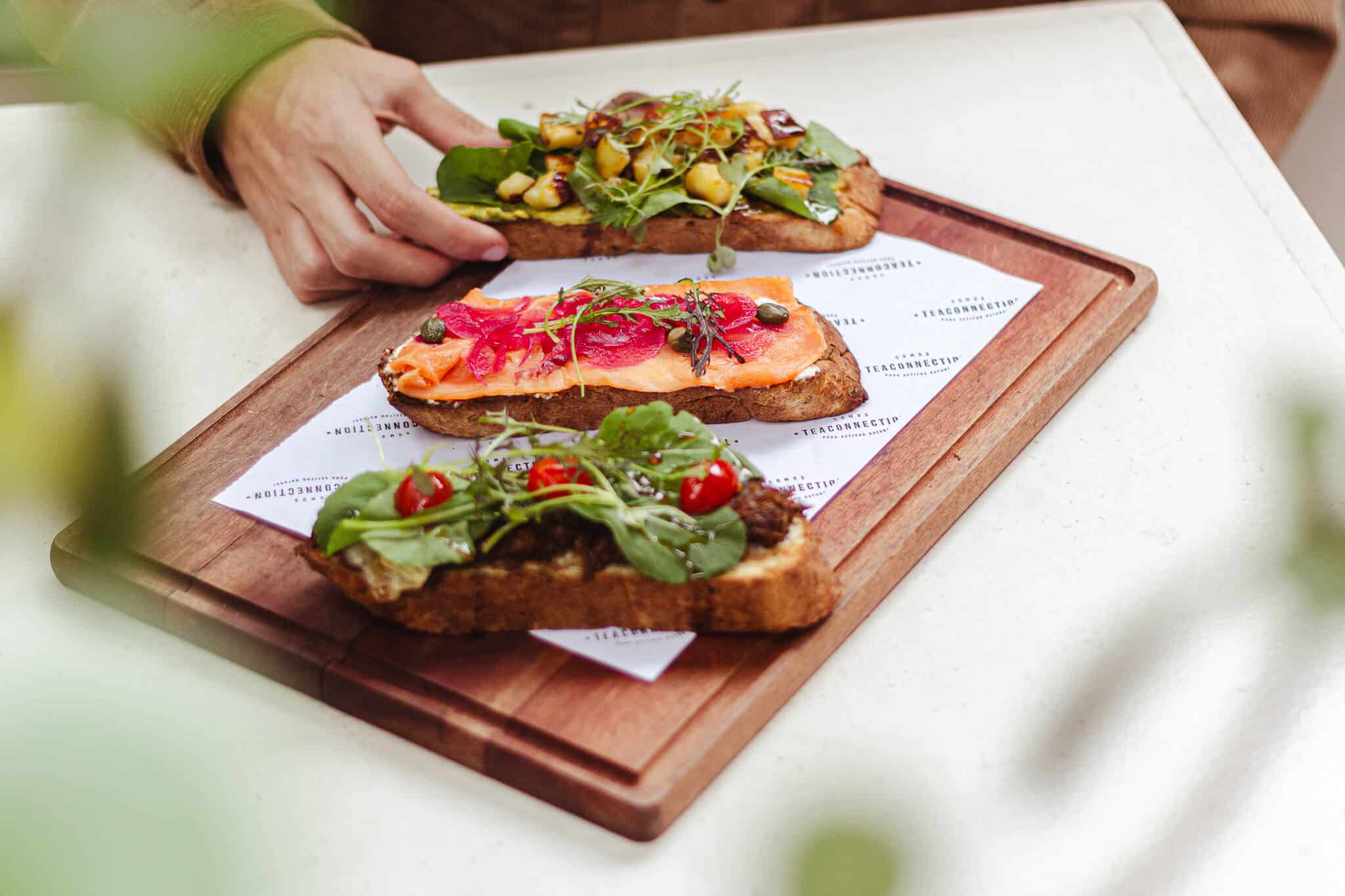 foto contem uma bandeja de madeira em cima de uma branca, contendo 3 toasts (torradas) do brunch do tea connection. Uma pessoa branca está segurando o toast de avocado. Em ordem: toast de vocado, salmão e carne.